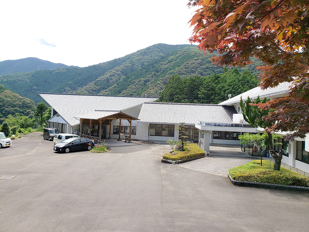 山間の介護施設の外観。モダンな白い建物と駐車場、秋の紅葉が映える。