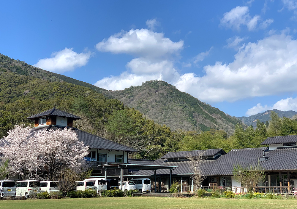 山々に囲まれた介護施設の外観。桜の木と広い敷地が特徴