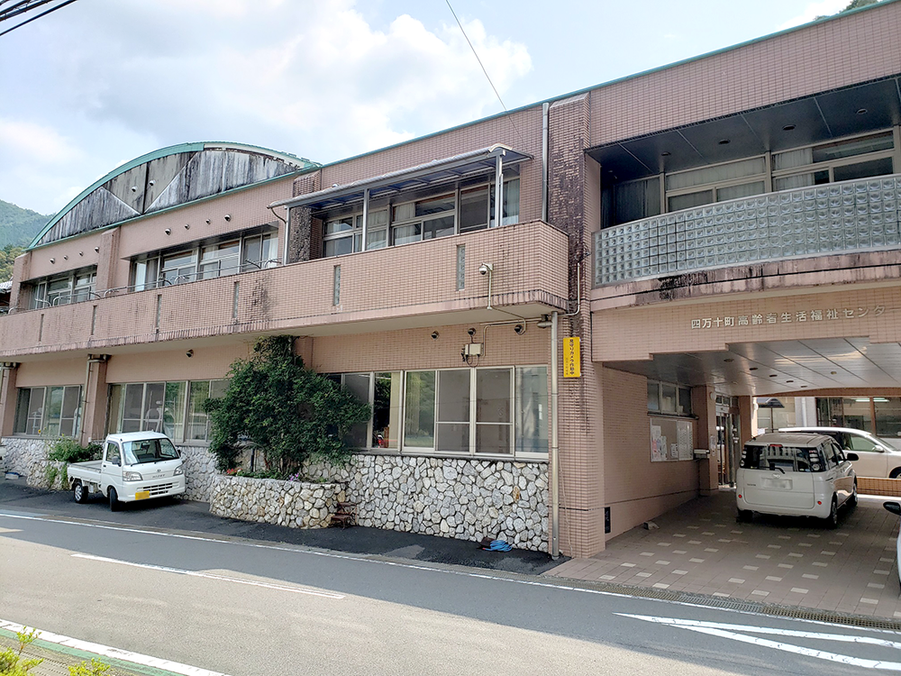 茶色レンガ造りの介護施設の外観。玄関前に駐車スペースがあり、山々に囲まれた立地。